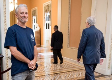 John Stewart standing in the hallways of congress semi-smiling in blue t-shirt